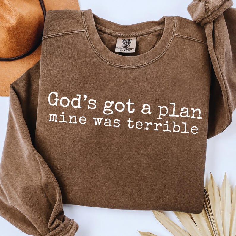 May include: A brown crewneck sweatshirt featuring the white printed text God's got a plan mine was terrible. The garment has a ribbed collar and long sleeves with a vintage-style wash. A label inside the neck reads Comfort Colors.