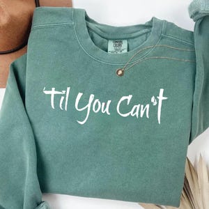 May include: A sage green crewneck sweatshirt featuring the white text 'til You Can't printed across the chest. The garment has a ribbed collar and a Comfort Colors brand label inside the neckline. A thin gold chain necklace rests on the fabric.