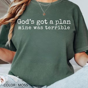 May include: A moss green short-sleeved t-shirt featuring the white text "God's got a plan mine was terrible" printed across the chest. The garment has a crew neckline and a relaxed fit, showcasing a solid colour fabric with a subtle textured appearance.