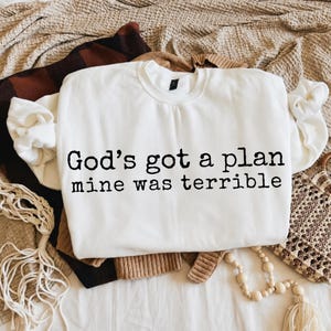May include: A white long-sleeve crewneck sweatshirt featuring black typewriter-style text printed on the front that reads, God's got a plan mine was terrible. The garment has a ribbed collar, cuffs, and hem, with a standard fit and long sleeves.