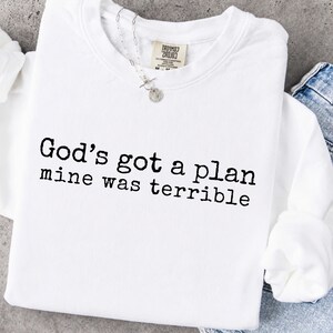 May include: A white long-sleeve crewneck sweatshirt featuring the black text "God's got a plan mine was terrible" printed in a typewriter-style font across the chest. The garment includes a tag at the inner neckline and a silver-toned chain necklace.