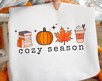 May include: White tote bag with a seasonal design. The design features a stack of books, an orange pumpkin, a red maple leaf, and a pumpkin spice latte. The words "cozy season" are printed below the illustrations.