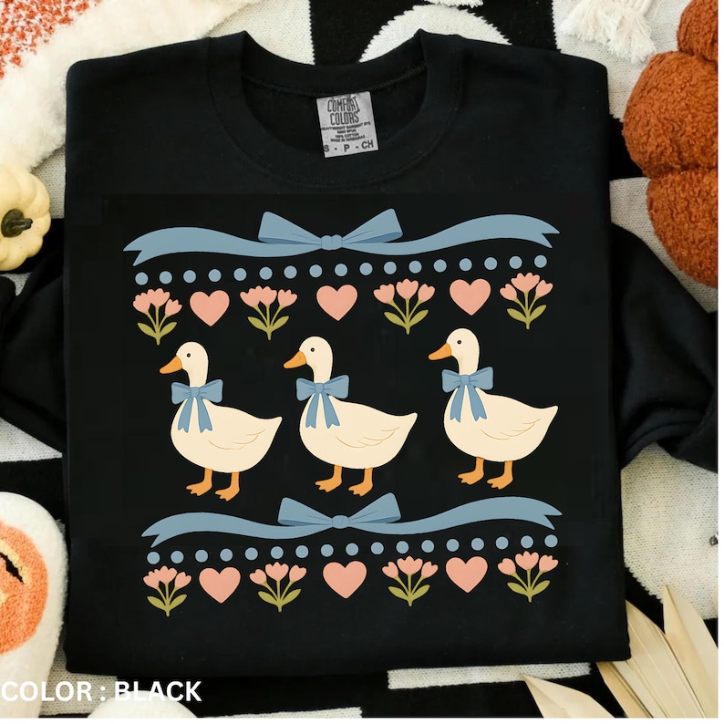Puede incluir: Una camiseta negra con un estampado gr&aacute;fico de tres patos de color crema con lazos azul claro. El dise&ntilde;o incluye filas de puntos azul claro, corazones rosas y peque&ntilde;as flores rosas, enmarcadas por dos cintas horizontales azul claro.