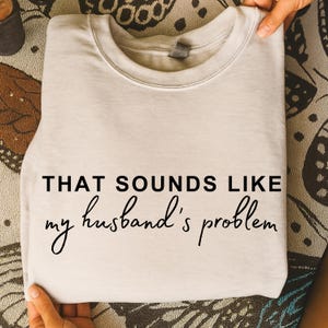 Pode incluir: Uma camisola bege clara dobrada com o texto "THAT SOUNDS LIKE my husband's problem" em escrita preta. A camisola é segurada por mãos, e o fundo tem um design estampado.