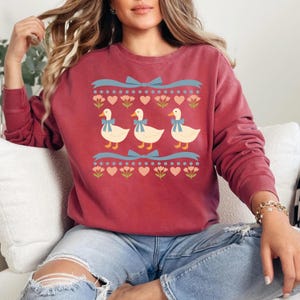 Puede incluir: Una sudadera de cuello redondo color rojo oscuro con un estampado gr&aacute;fico de tres patos blancos con lazos azules. El dise&ntilde;o incluye filas de cintas azul claro, corazones rosas y peque&ntilde;as flores verdes sobre y debajo de los patos.