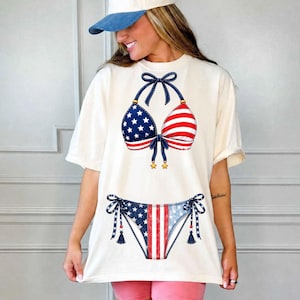 Peut inclure: T-shirt crème avec un motif de bikini patriotique. Le haut présente une coupe bleue parsemée d'étoiles et une coupe à rayures rouges et blanches. Le bas a un motif assorti. Le t-shirt est de coupe décontractée.