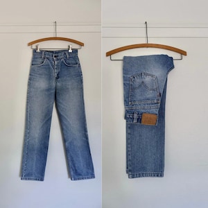 May include: Two pairs of light-wash denim jeans hanging on wooden hangers. One pair is displayed fully, while the other is folded. The jeans have a classic five-pocket design and a button and zipper closure.