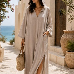 Pode incluir: Um vestido caftan bege com capuz, mangas compridas, decote em V profundo e uma fenda lateral. A peça inclui bolsos laterais e é combinada com uma bolsa de palha trançada e sandálias castanhas. O texto Shahig Fabric Fusion é visível.