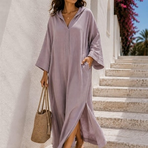 May include: A long, loose-fitting lavender hooded dress with wide three-quarter length sleeves, a V-neckline, and side slits. The dress features side pockets and is paired with brown leather sandals and a woven straw tote bag. Text reads Shahig Fabric Fusion.