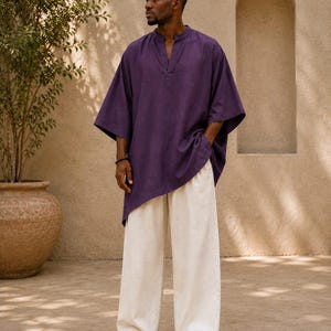 Puede incluir: Una túnica de color morado intenso con cuello en V y mangas anchas hasta el codo, combinada con pantalones holgados de color blanco roto. El conjunto presenta una textura de tela y el texto Shahiq Fabric Fusion en la parte inferior.