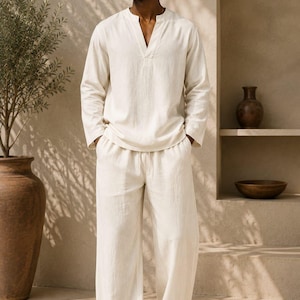 May include: A cream-colored long-sleeve shirt with a V-neckline and matching loose-fitting trousers with an elastic drawstring waist. The set is made of a textured fabric and is paired with black slide sandals and dark-tinted sunglasses.