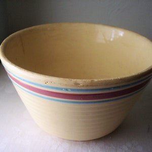 May include: A vintage, cream-colored mixing bowl with a wide, rounded shape. The bowl features two horizontal stripes: a central magenta stripe bordered by thinner light blue stripes. The rim shows slight wear, adding to its antique charm.