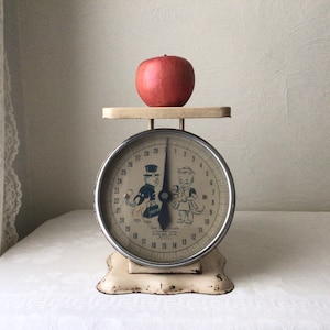May include: Vintage white kitchen scale with a black and white illustration of a cat and a man wearing a top hat. The scale has a red apple on the platform and the face reads "Jay's Scale".