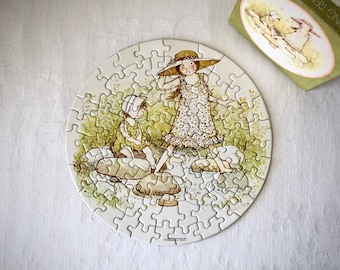 May include: A round jigsaw puzzle with a white background and a colorful image of two children playing in a forest. The girl is wearing a white hat and a floral dress, and the boy is wearing a blue shirt and brown pants. The puzzle is surrounded by a white border.