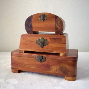 May include: Three wooden boxes stacked on top of each other. The boxes are made of cedar wood and have brass hinges and latches. The top box is oval shaped, the middle box is rectangular, and the bottom box is rectangular with rounded edges.