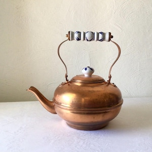 May include: A copper tea kettle with a round body, curved spout, and a handle with white and blue ceramic accents. The lid features a matching ceramic knob. The kettle is set against a neutral background.