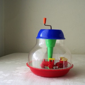 May include: A red and yellow toy train spins inside a clear plastic dome with a blue lid and a green spinning top. The toy is powered by a hand crank.