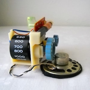 May include: Close-up of a vintage radio tuner with a dial that reads from 530 to 1200. The tuner is attached to a small speaker with a black cloth covering.