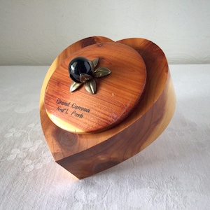 May include: A heart-shaped wooden box with a lid. The lid has a black glass ball with a gold metal flower design. The box is inscribed with "Grand Canyon Nat'l Park".