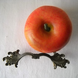 May include: A decorative metal drawer pull with ornate scrollwork on each end. The pull is in a dark bronze finish and is resting on a red apple.