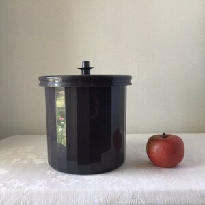 May include: A black plastic ice bucket with a lid and a small handle on top. The bucket is sitting on a white tablecloth with a red apple next to it.