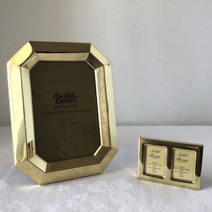 May include: Two gold-toned picture frames. The larger frame is octagonal and has the text "Solid Brass Photo Frame hand polished & lacquer coated 5 x 7 13cm x 18cm". The smaller frame is rectangular and has two openings for photos. Both frames have the text "Solid Brass".