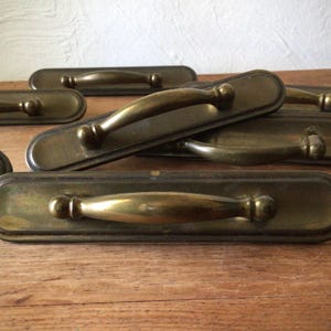 May include: A collection of antique brass drawer pulls. Each pull features a rectangular backplate with rounded ends and a curved handle. The brass has a warm, aged patina, suggesting vintage or reclaimed hardware. The pulls are arranged on a wooden surface.
