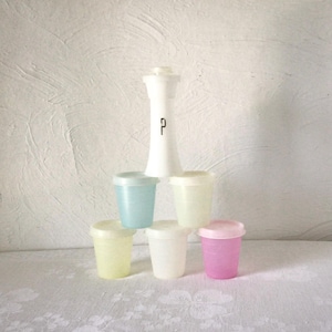 May include: A white pepper mill with a black letter P printed on the front, stacked above five small, translucent plastic containers with lids in light blue, pale yellow, off-white, cream, and pink, arranged in a pyramid formation.
