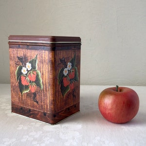 May include: A square, brown metal tin with a woodgrain pattern and painted images of strawberries and white flowers. The tin has a white rim around the top.