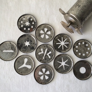 May include: A set of 10 metal cookie cutters with different shapes, including stars, flowers, and geometric designs. The cutters are arranged in a circle on a white surface.