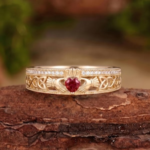 May include: A gold ring featuring a central heart-shaped red gemstone held by two hands, topped with a small crown. The band displays intricate Celtic knotwork patterns on both sides and is accented with a row of small clear stones along the top edge.