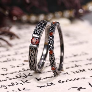 Könnte beinhalten: Zwei Ringe aus schwarzem Metall mit komplizierten keltischen Knotenmustern. Der breitere Ring ist mit einem einzelnen runden, tiefroten Granat-Edelstein besetzt, während das dünnere, stapelbare Band mit mehreren kleinen, runden roten Steinen verziert ist.
