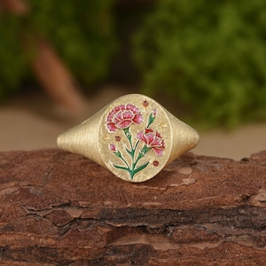 May include: A gold signet ring with a brushed matte finish featuring an oval face. The center displays an enamel design of two pink carnations with green stems and leaves, accented by small engraved stars and tiny red circular stone inlays.