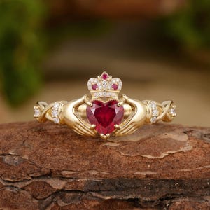 May include: A gold Claddagh ring featuring a central heart-shaped red gemstone held by two hands. The ring includes a crown above the heart, accented with small white stones and a red stone, with additional white stones along the band.