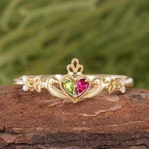 May include: A gold Claddagh ring featuring a central heart-shaped peridot and a heart-shaped ruby, topped with a small crown. The band displays intricate metalwork details and is accented with two small clear stones on each side of the center.