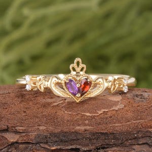 May include: A gold Claddagh ring featuring a central heart-shaped purple amethyst and a red garnet stone, topped with a crown. The band includes intricate metalwork details and small clear accent stones on each side of the central heart design.