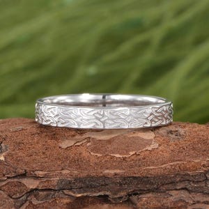 Könnte beinhalten: Ein silberner Ring mit einem eingravierten Muster. Der Ring befindet sich auf einer braunen, strukturierten Oberfläche mit einem verschwommenen grünen Hintergrund. Das Design des Rings weist ein sich wiederholendes, wirbelndes Muster um das Band auf.