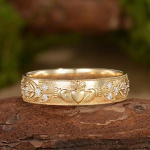 May include: A gold Claddagh ring featuring a central heart held by two hands topped with a crown. The band is decorated with intricate engraved patterns and small, clear, round-cut stones set along the sides of the central motif.