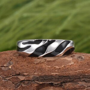 May include: A silver ring with a black enamel wave design. The ring is set against a natural wood surface with a blurred green background.