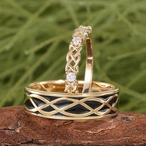 Könnte beinhalten: Zwei goldene Eheringe. Ein Ring hat ein schwarzes Emaille-Design mit einem goldenen Rand. Der andere ist ein dünner Goldring mit einem keltischen Knotenmuster und kleinen Diamanten.