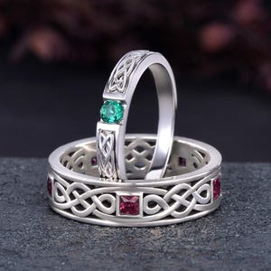 May include: Two silver rings with Celtic knot designs. One ring features a green gemstone, while the other has red gemstones set within the knotwork. The rings are displayed on a dark, textured surface.