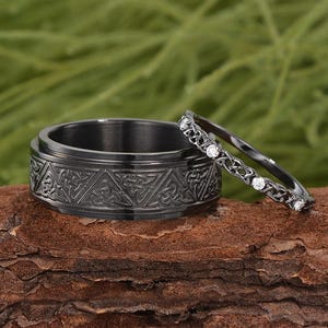 Könnte beinhalten: Zwei schwarze Ringe. Einer ist breit mit einem keltischen Knotenmuster. Der andere ist ein dünnes Band mit kleinen, klaren Steinen. Die Ringe werden auf einem Stück braunem Holz mit verschwommenem grünem Hintergrund präsentiert.