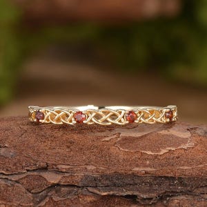 Könnte beinhalten: Ein goldener Ring mit einem komplexen, geflochtenen Metallmuster entlang der Ringschiene, akzentuiert durch vier kleine, runde, tiefrote Edelsteine, die gleichmäßig verteilt sind und ein durchgehendes dekoratives Motiv auf der Oberfläche bilden.