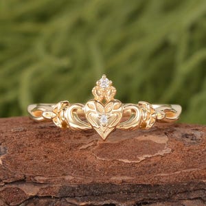 Könnte beinhalten: Ein goldener Claddagh-Ring mit einem zentralen Herzmotiv, das von einer Krone gekrönt und von zwei Händen flankiert wird. Der Ring ist mit zwei kleinen, klaren, rund geschliffenen Edelsteinen besetzt, einer im Herzen und einer an der Krone.