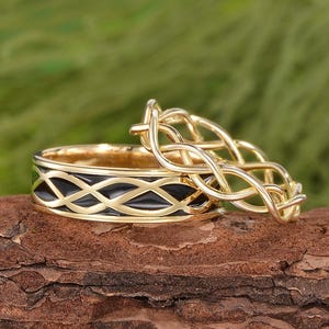 Könnte beinhalten: Zwei goldfarbene Ringe mit aufwendigen keltischen Knotenmustern. Ein Ring hat eine schwarze Emaille-Einlage mit sich wiederholendem Muster. Der andere Ring ist ein filigranes Design. Die Ringe werden auf einem Holzstück präsentiert.