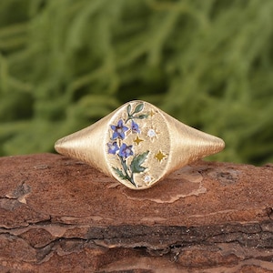 May include: Gold signet ring with an oval face featuring a floral design. The ring's face showcases blue flowers, green leaves, and small star and diamond accents. The ring has a textured band and a warm, golden hue.