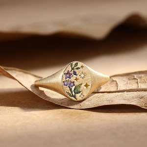 K&ouml;nnte beinhalten: Ein goldener Siegelring mit geb&uuml;rstetem, mattem Finish und ovaler Ringplatte. Die Platte ist mit einem floralen Emaille-Design in Blau- und Gr&uuml;nt&ouml;nen verziert, akzentuiert durch kleine eingravierte Sternformen und zwei winzige klare Steine.