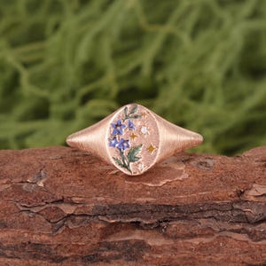 K&ouml;nnte beinhalten: Ein Siegelring aus Ros&eacute;gold mit strukturiertem Ringband und flacher, ovaler Oberseite. Die Oberseite ist mit einem Emaille-Design aus blauen Blumen mit gr&uuml;nen Bl&auml;ttern verziert, akzentuiert durch kleine goldfarbene Sterne und zwei klare Steine.