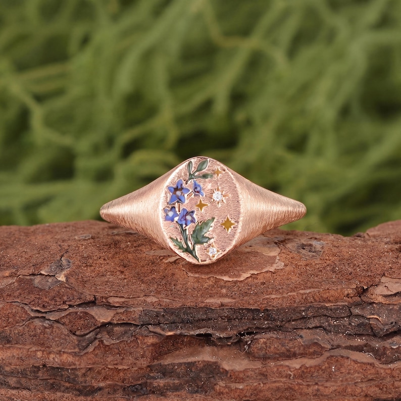 K&ouml;nnte beinhalten: Ein Siegelring aus Ros&eacute;gold mit strukturiertem Ringband und flacher, ovaler Oberseite. Die Oberseite ist mit einem Emaille-Design aus blauen Blumen mit gr&uuml;nen Bl&auml;ttern verziert, akzentuiert durch kleine goldfarbene Sterne und zwei klare Steine.