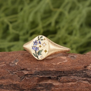 K&ouml;nnte beinhalten: Ein goldener Siegelring mit geb&uuml;rsteter Textur und ovaler Ringplatte. Die Platte zeigt ein Emaille-Design aus blauen Blumen mit gr&uuml;nen Bl&auml;ttern, akzentuiert durch kleine goldene Sterne und zwei winzige, in die Metalloberfl&auml;che eingelassene klare Steine.